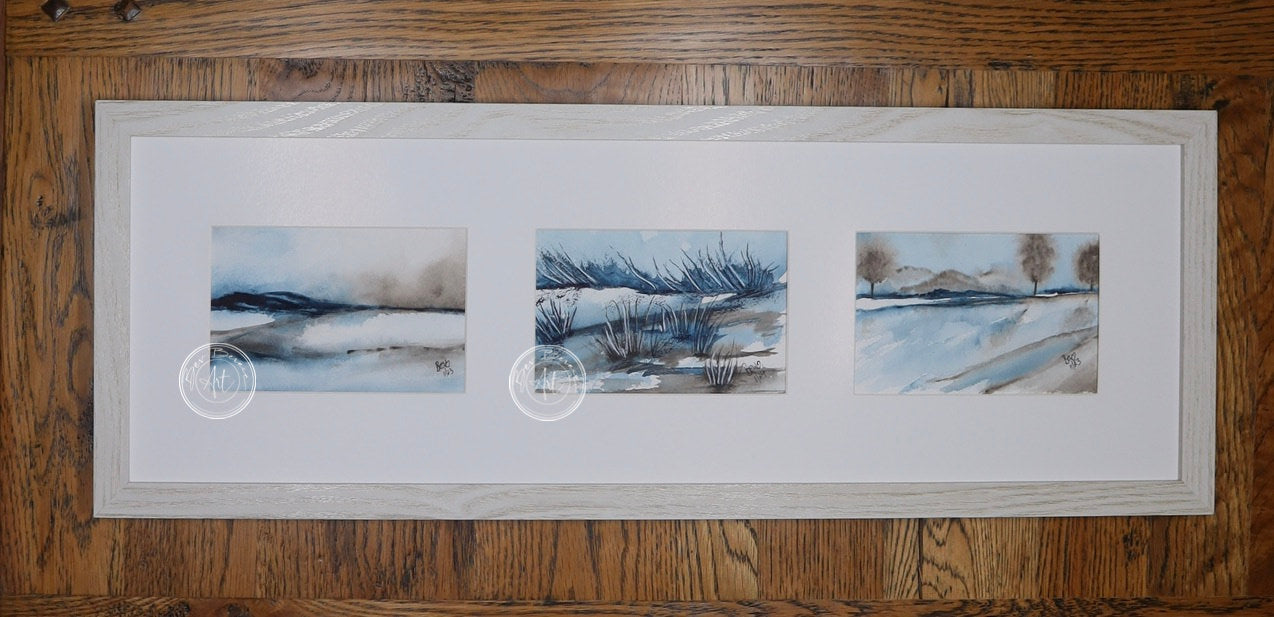 Three small square watercolor paintings in a row, depicting a minimalist winter landscape with blue, gray, and brown tones. The paintings are framed in a light wood frame with a wide white mat. Each painting shows a different aspect of the landscape, including water, reeds, and distant trees.