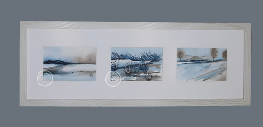 Three small square watercolor paintings in a row, depicting a minimalist winter landscape with blue, gray, and brown tones. The paintings are framed in a light wood frame with a wide white mat. Each painting shows a different aspect of the landscape, including water, reeds, and distant trees.