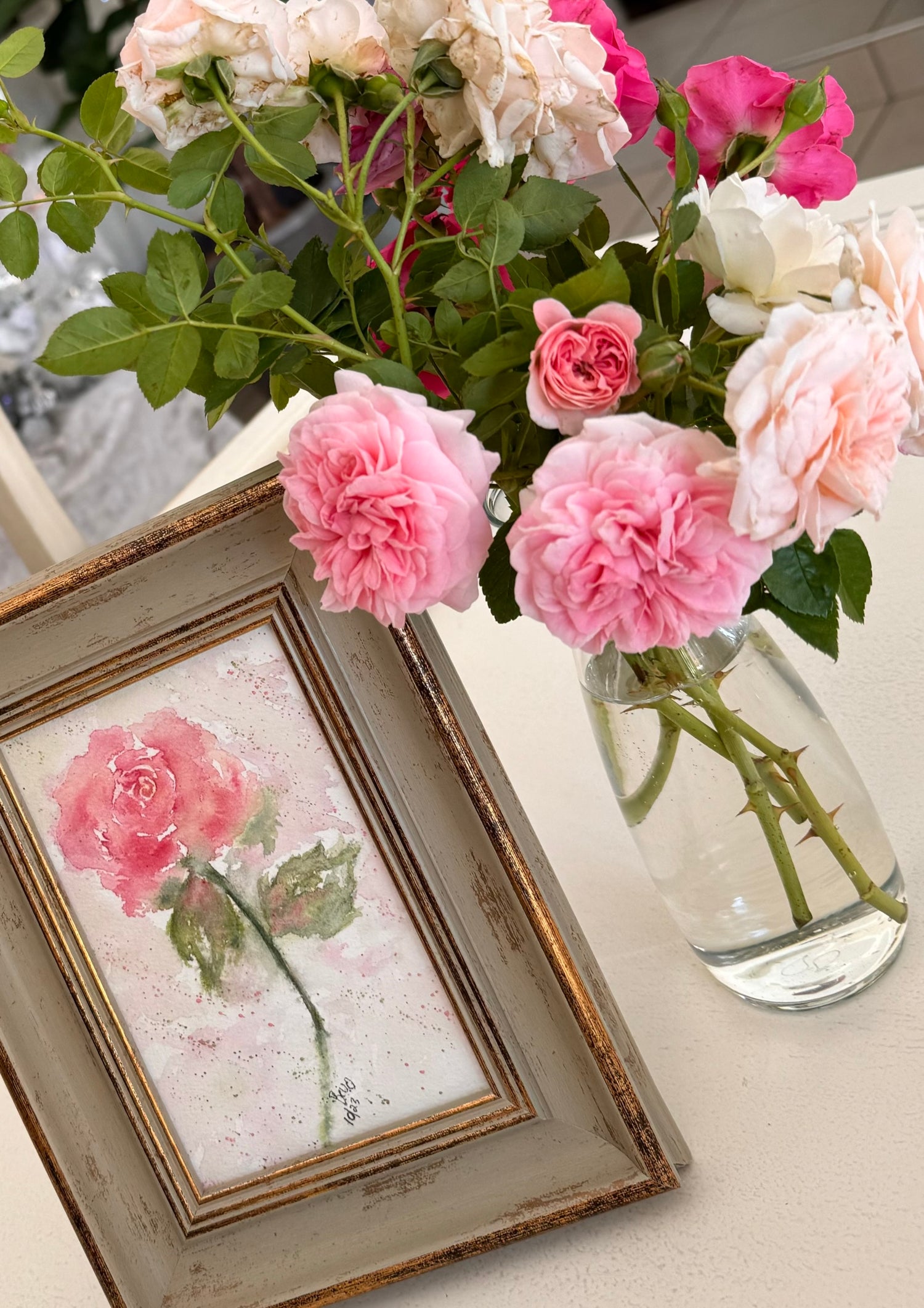 A framed watercolour painting of a pink rose displayed alongside a glass vase of fresh pink roses and green foliage, creating a harmonious floral arrangement.