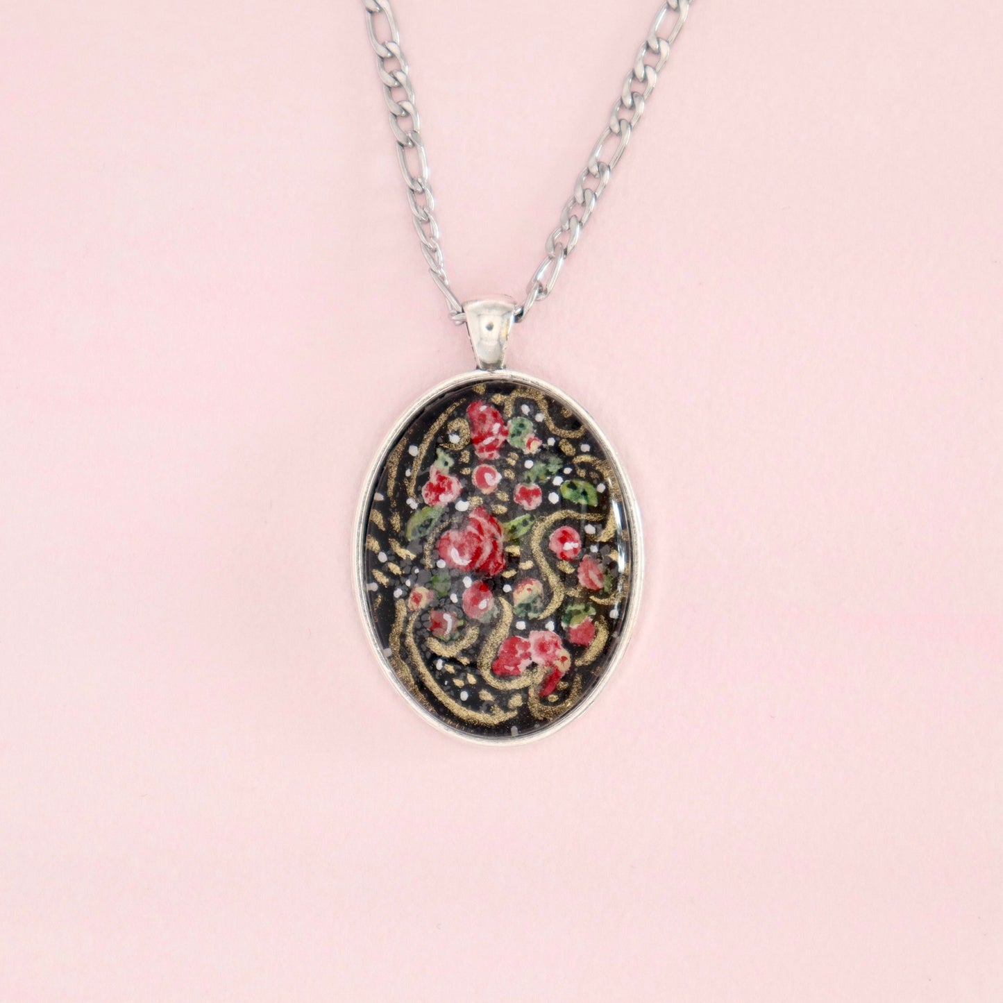A close-up of the Midnight Bloom No. 1 hand-painted miniature watercolor pendant, featuring pink flowers with gold accents on a black background, encased in an oval silver-tone setting on a silver chain.