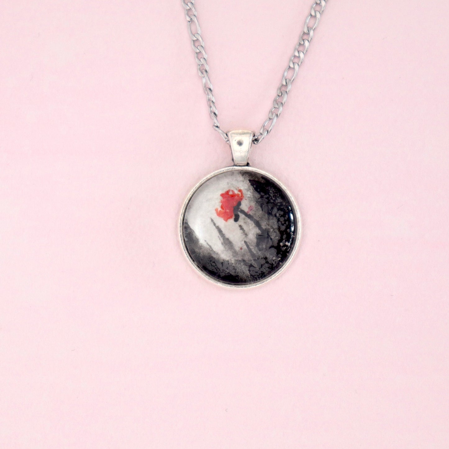 A silver necklace with a round pendant. The pendant features an abstract painting with a red flower against a black and white background, possibly depicting a snowy landscape.
