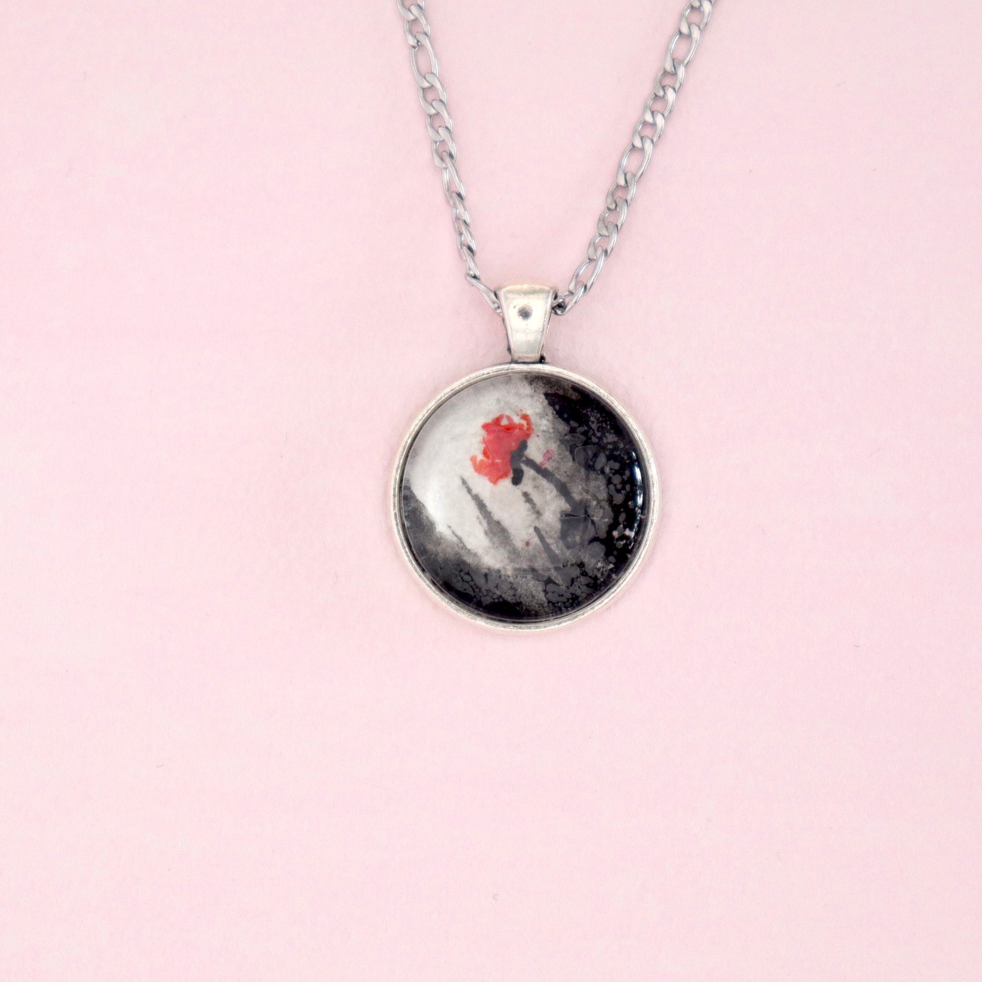 A silver necklace with a round pendant. The pendant features an abstract painting with a red flower against a black and white background, possibly depicting a snowy landscape.