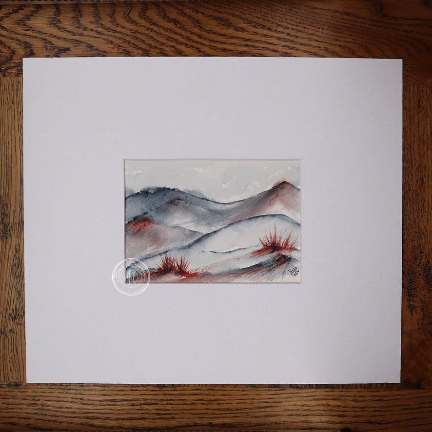Watercolour painting of a rolling landscape with blue-grey hills and splashes of red, possibly representing desert shrubs, in the foreground.